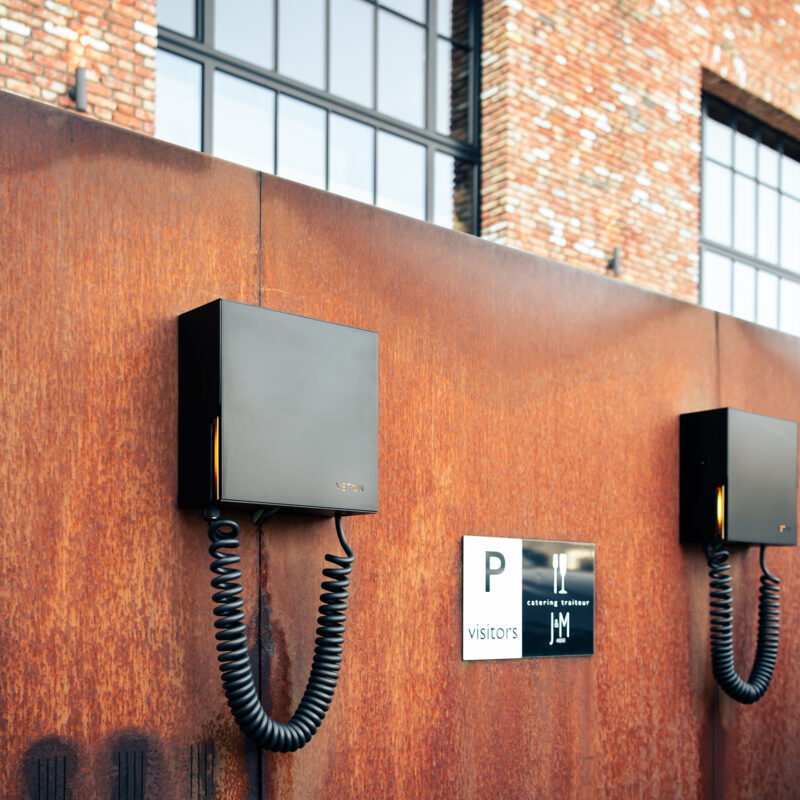 Two Veton Wall Plus chargers on a corten steel facade at the entrance, visitor signage alongside.